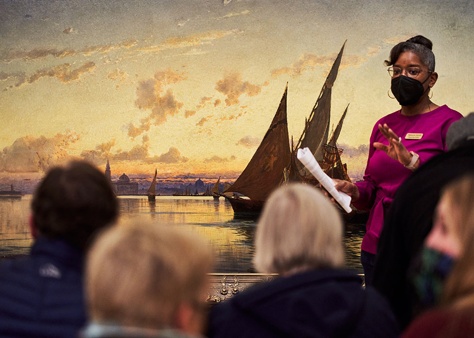 A person addressing a group of people while standing in front of an artwork of a harbor at sunset